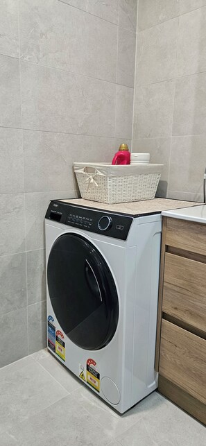 A front-loading washing machine in a kitchen with grey tiled walls and wooden cabinets. - Spacious Holiday Home in Epping, 3BR, 2 baths, Sleeps 6, Free Parking. (Epping)