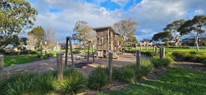 A playground with a wooden climbing structure and swings, surrounded by grass and trees.