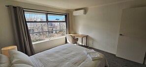A hotel room with a bed, a desk, a chair, a window with curtains, and an air conditioning unit. - Spacious Holiday Home in Epping, 3BR, 2 baths, Sleeps 6, Free Parking. (Epping)