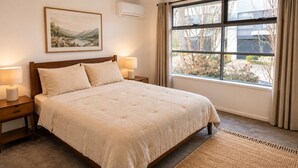 Een slaapkamer met een groot bed, twee nachtkastjes met lampen, een raam met gordijnen, en een lijst met een afbeelding aan de muur.