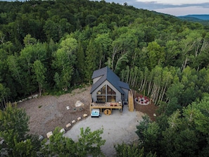A cabin surrounded by dense forest, with a patio area and outdoor seating. - Close to Sunday River: Hot Tub, Sauna, Cold Plunge (Bethel)