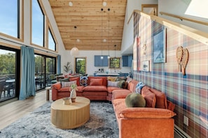 A spacious living area with a large sofa, a round coffee table, and a view of the outdoors through large windows. - Close to Sunday River: Hot Tub, Sauna, Cold Plunge (Bethel)