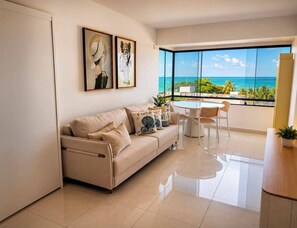 A modern living room with a beige sofa, a round dining table, and chairs. There are two framed artworks on the wall, and a large window offering a view of the ocean. - Ocean View Ponta Verde (Maceió)
