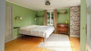 Ein Schlafzimmer mit Bett, Nachttischlampe, Kommode, Fenster mit Vorhängen und einer Wand mit einer Pflanzenablage.