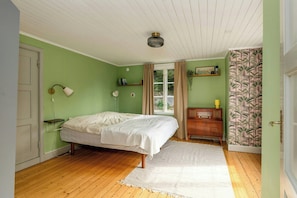 Ein Schlafzimmer mit Bett, Nachttischlampe, Kommode, Fenster mit Vorhängen und einer Wand mit einer Pflanzenablage.