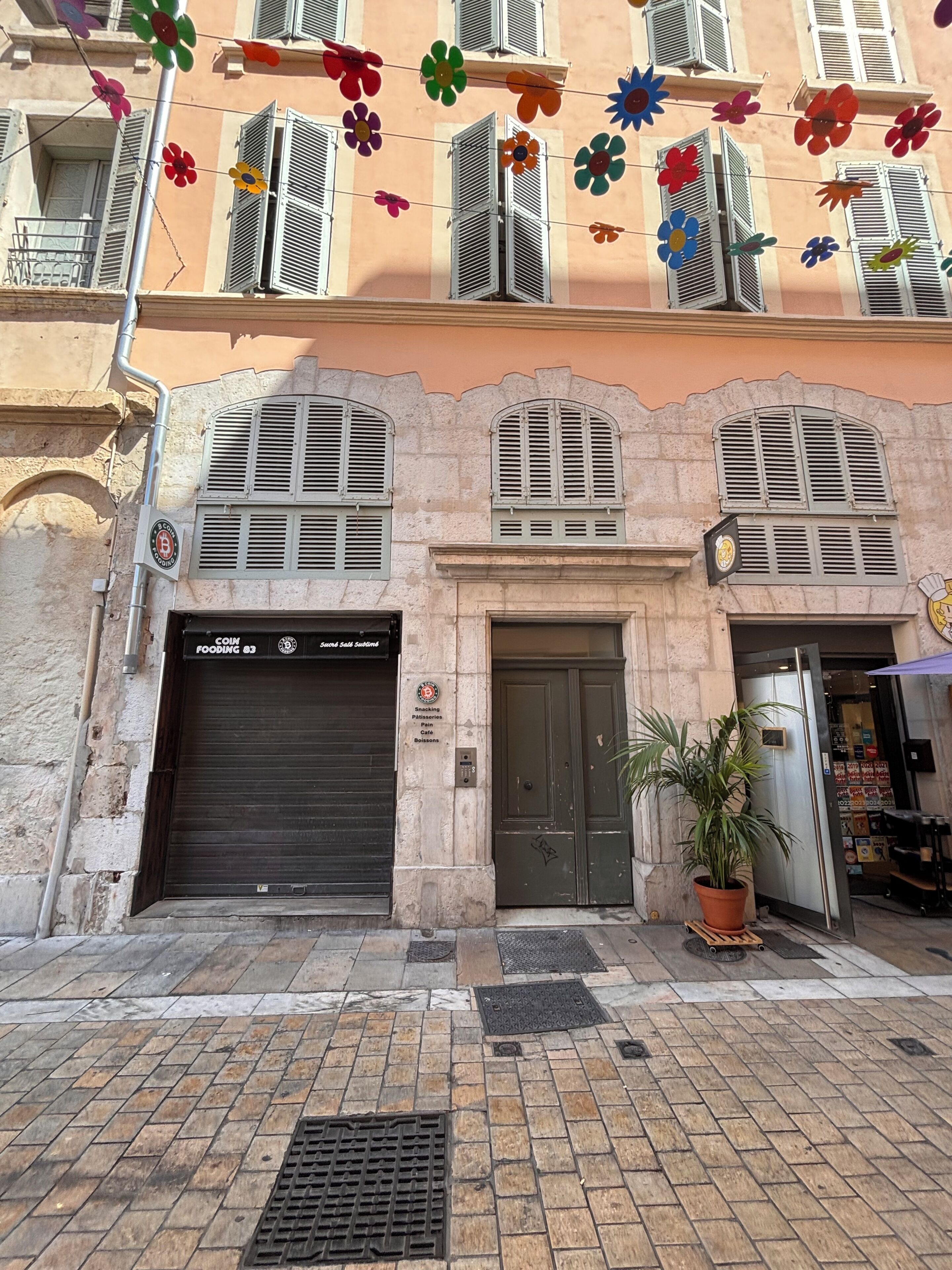 Un bâtiment dont l’entrée est fermée par un volet, avec un pot de fleurs et une enseigne indiquant qu’il s’agit d’un point de vente de nourriture fonctionnant avec des pièces de monnaie.