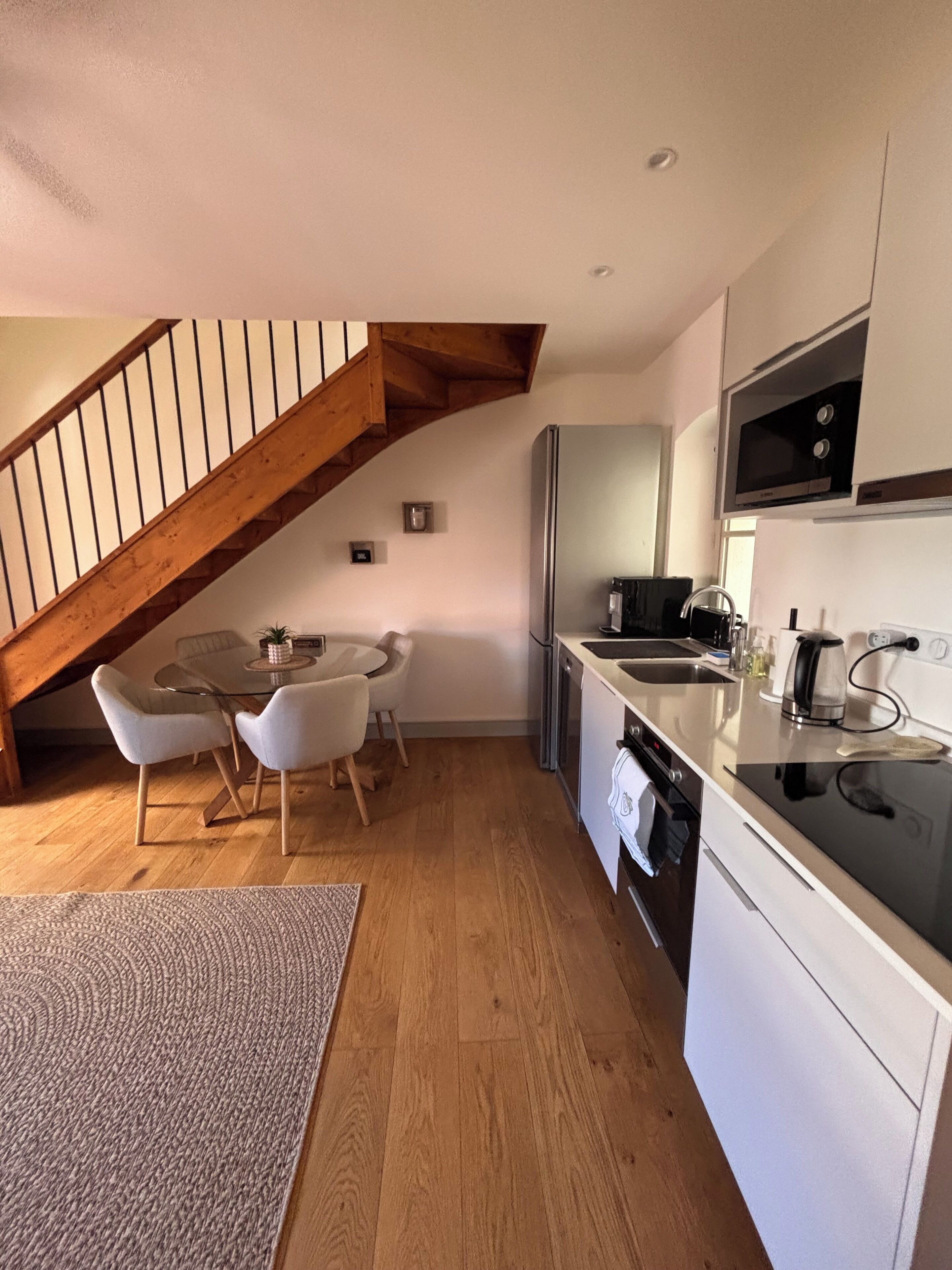 A modern kitchen with a wooden staircase, a dining area with a glass table and chairs, and a built-in oven.