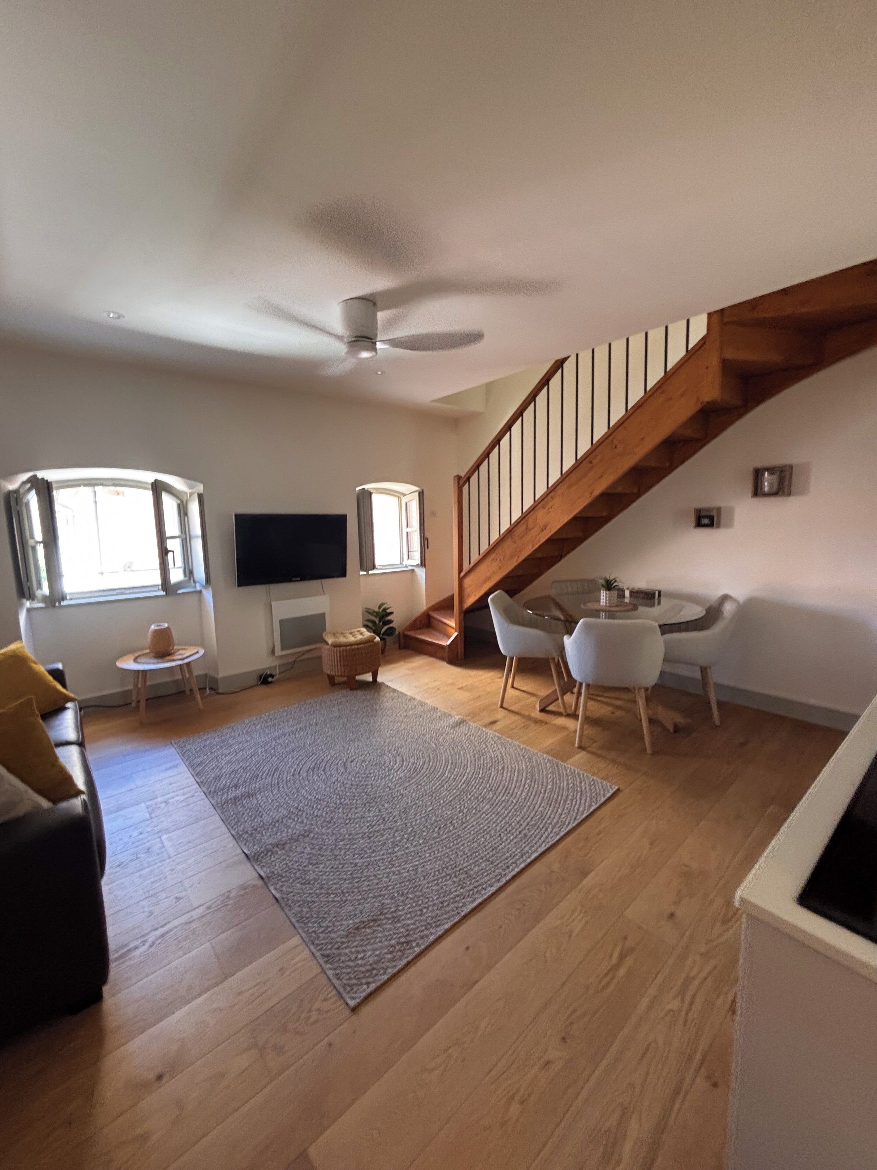 A spacious living area with a wooden staircase, a dining table with chairs, a sofa, and a television.