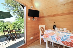 A wooden cabin with a TV mounted on the wall, a table with a floral tablecloth, and a chair. - Ivy Tor (Tavistock)