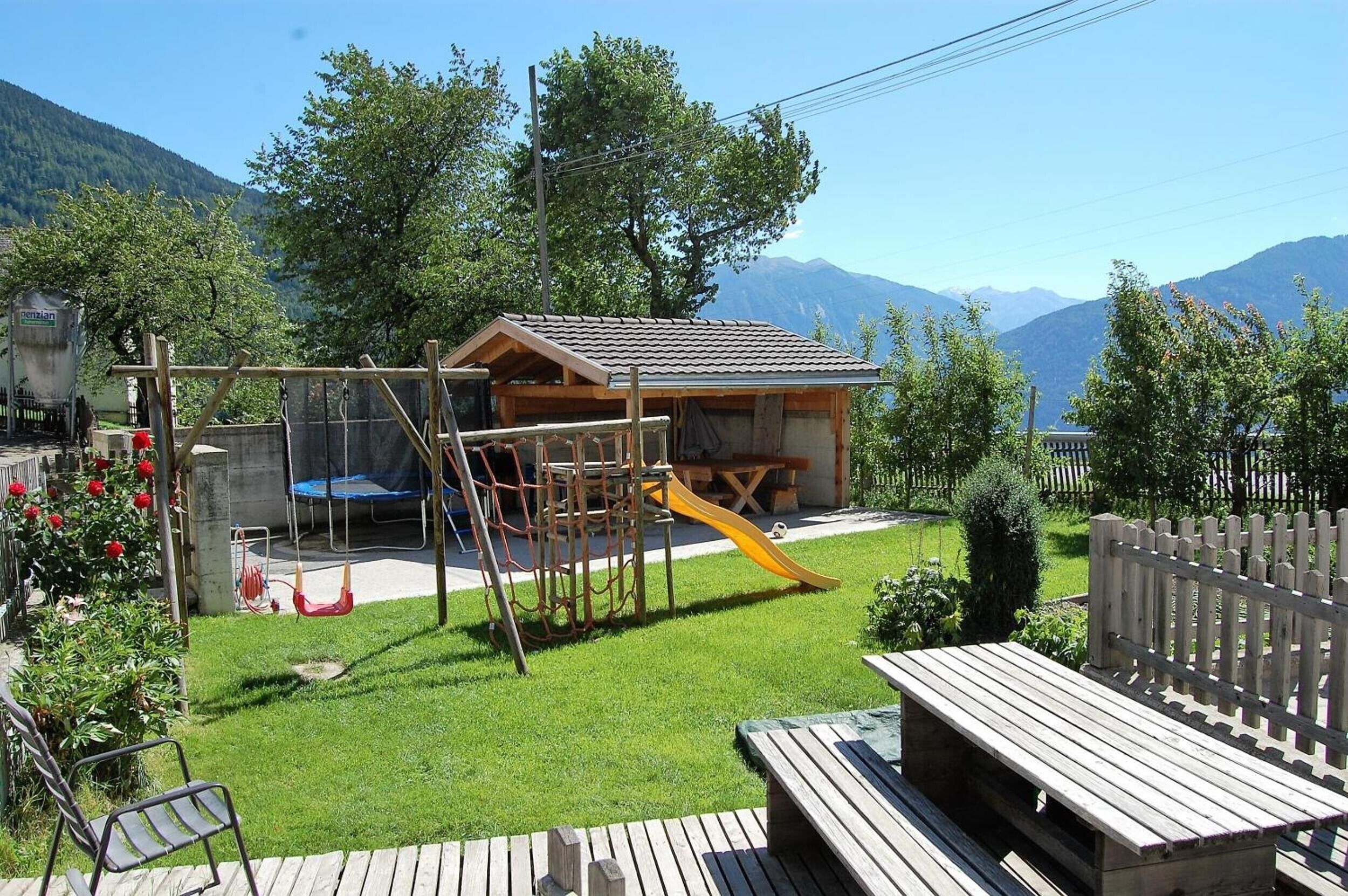 A wooden play structure with a slide, a picnic table, and a bench in a backyard with a mountain backdrop.