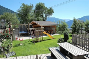 Une structure de jeu en bois comprenant un toboggan, une table de pique-nique et un banc, installée dans un jardin avec une vue sur les montagnes en arrière-plan.