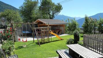 A wooden play structure with a slide, a picnic table, and a bench in a backyard with a mountain backdrop.