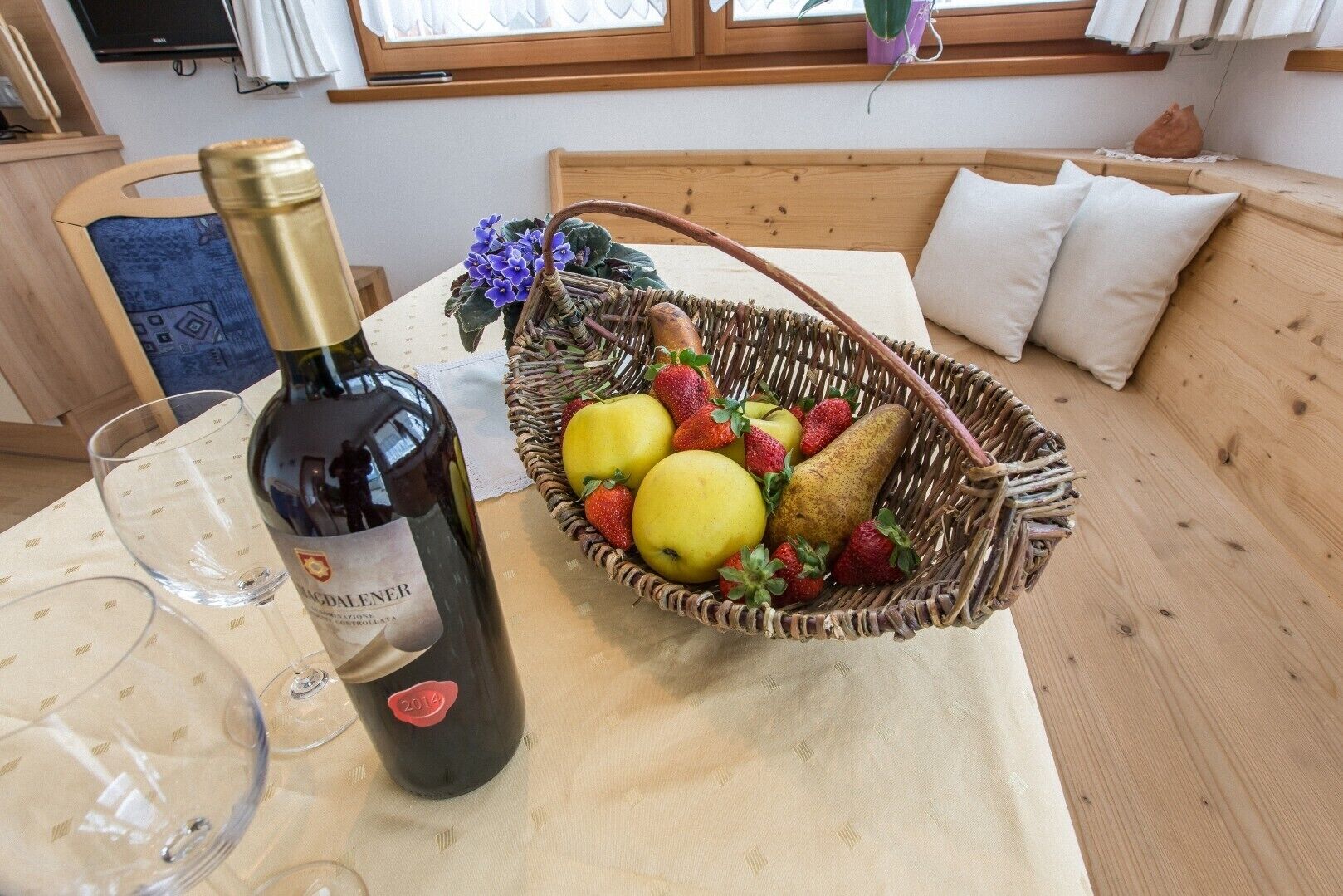 Een fles wijn, een glas en een mand met fruit op een tafel in een kamer met een houten bank en kussens.