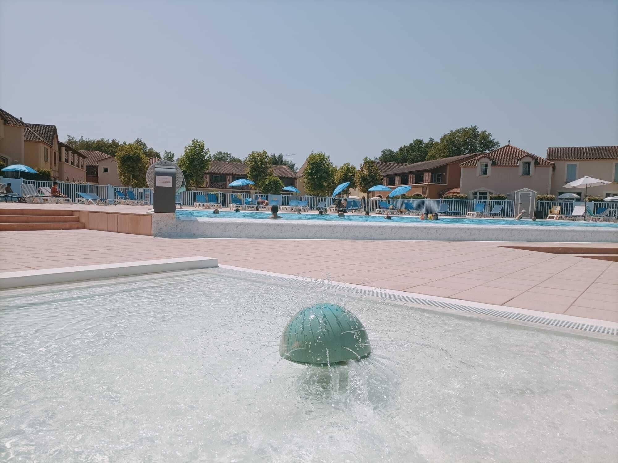 A swimming pool area with a water fountain feature, lounge chairs, and umbrellas.