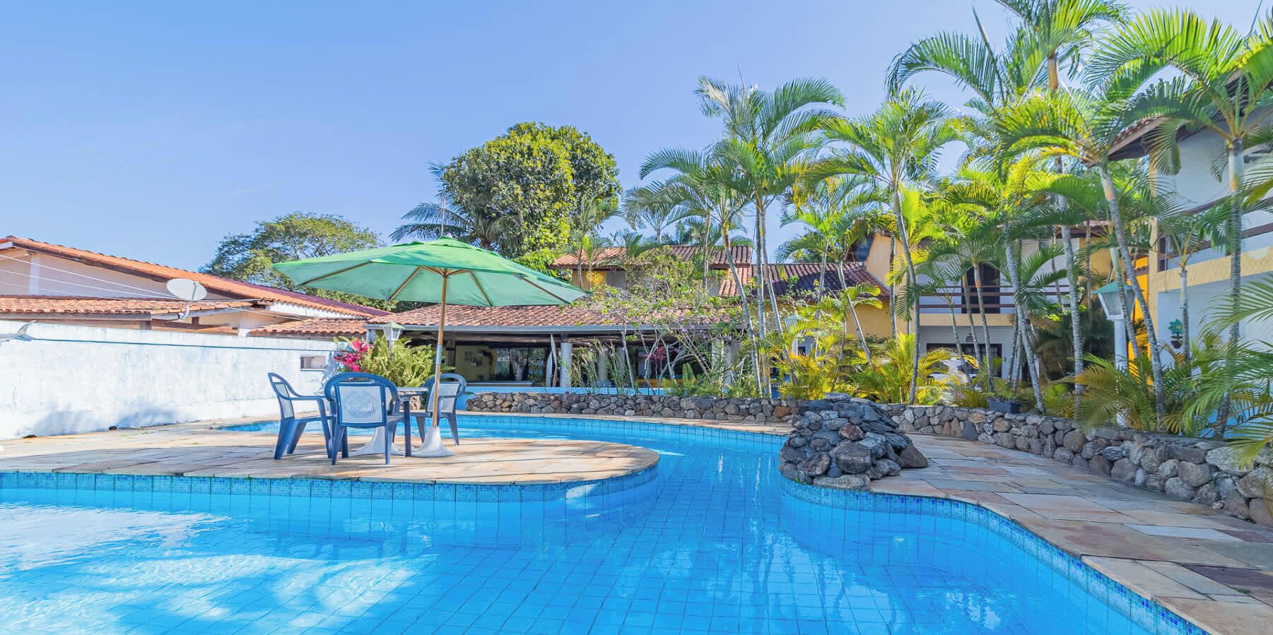A swimming pool with a stone border, surrounded by palm trees and a patio area with chairs and an umbrella.