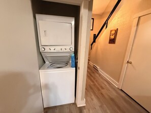 A compact laundry room with a front-loading washer and dryer, a blue detergent bottle, and a wooden floor. - Poconos Ski-In Chalet on Telstar | Loft, Private Deck & Gas Grill (Lake Harmony)