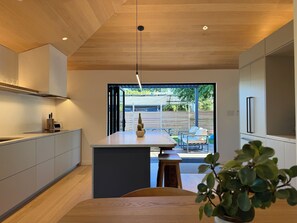 A modern kitchen with a wooden ceiling, a central island with seating, and a view to an outdoor patio. - Alki Beach Hygge Home- Modern 2 Bedroom with Backyard Oasis, Hot Tub (Seattle)