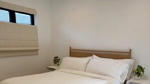 Uma cama bem arrumada com estrutura de madeira e roupa de cama branca, uma pequena mesa de cabeceira com uma planta e uma janela com persianas.