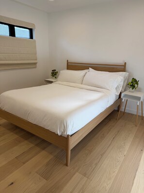 Ein ordentlich bezogenes Bett mit Holzrahmen und weißer Bettwäsche, ein kleiner Nachttisch mit Pflanze und ein Fenster mit Jalousien.