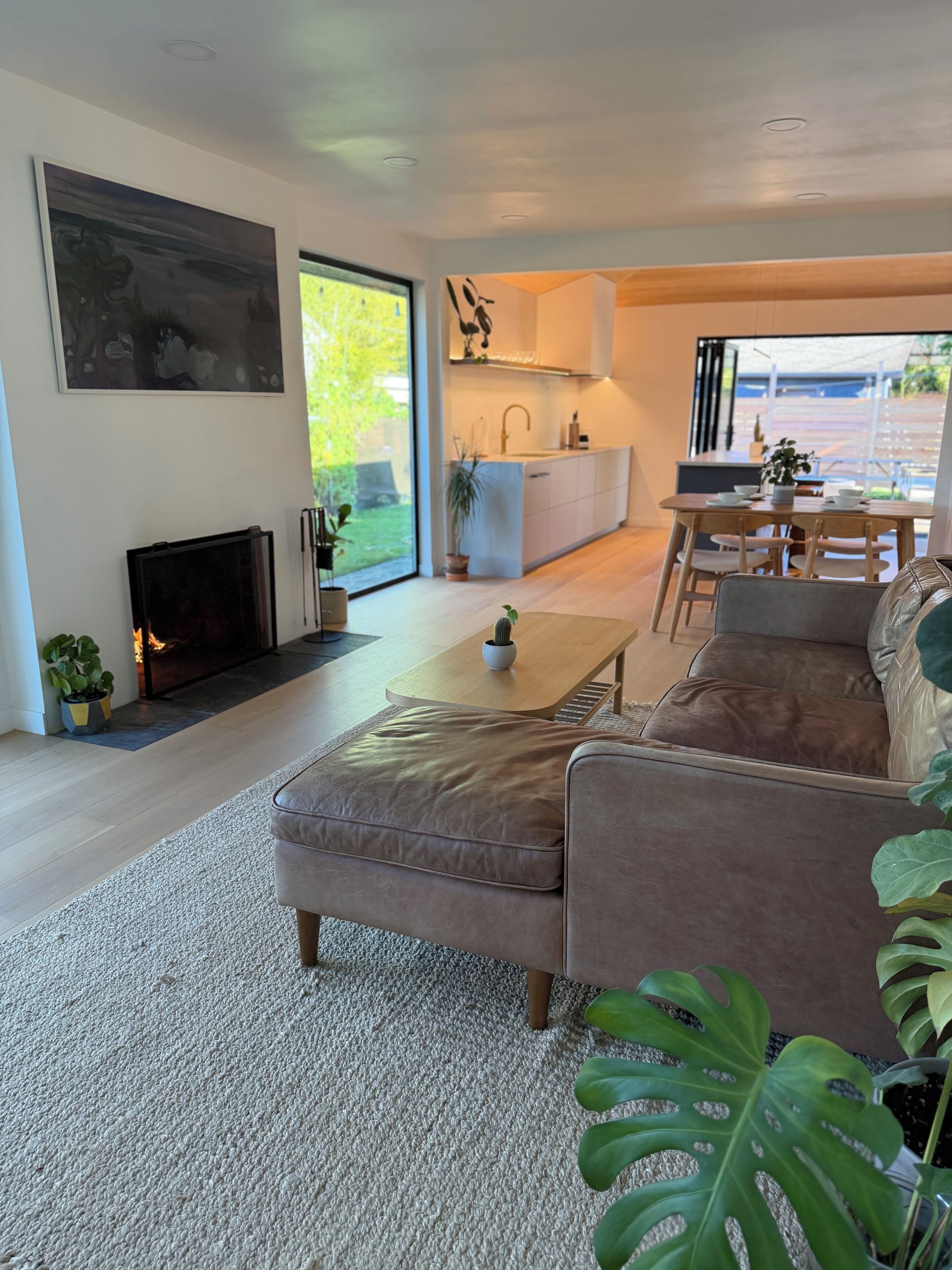 A modern living room with a fireplace, a sofa, a coffee table, and a view of the kitchen and outdoor area.