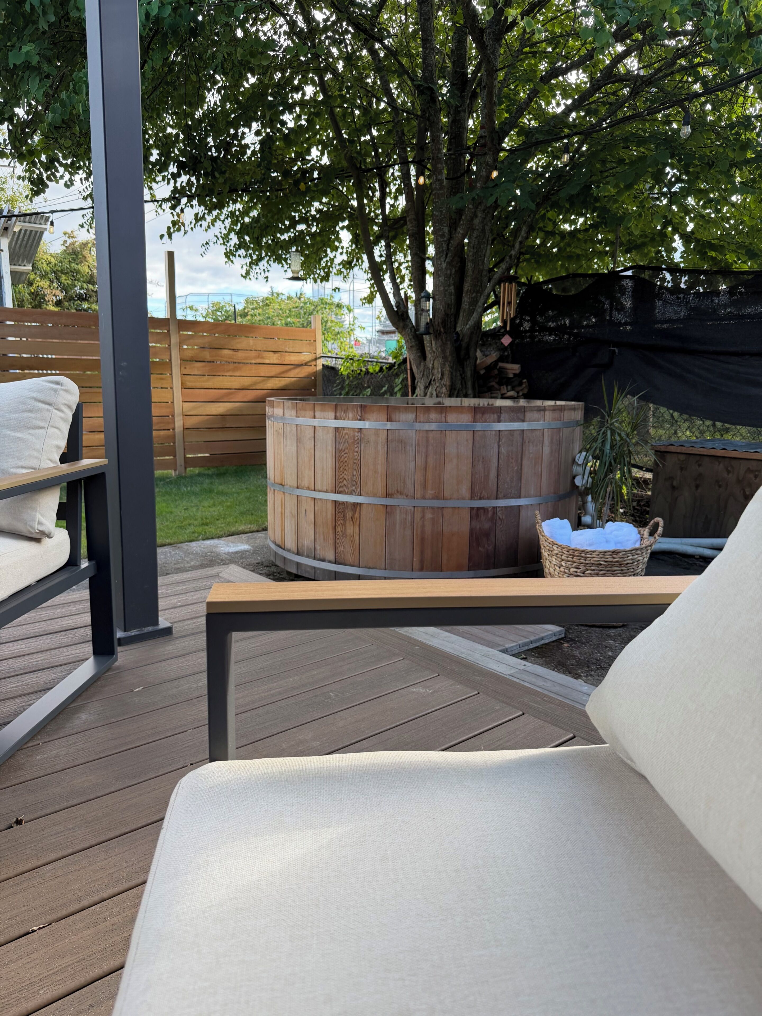 Een houten terras met een rond houten bad, een rieten mand en een bank.
