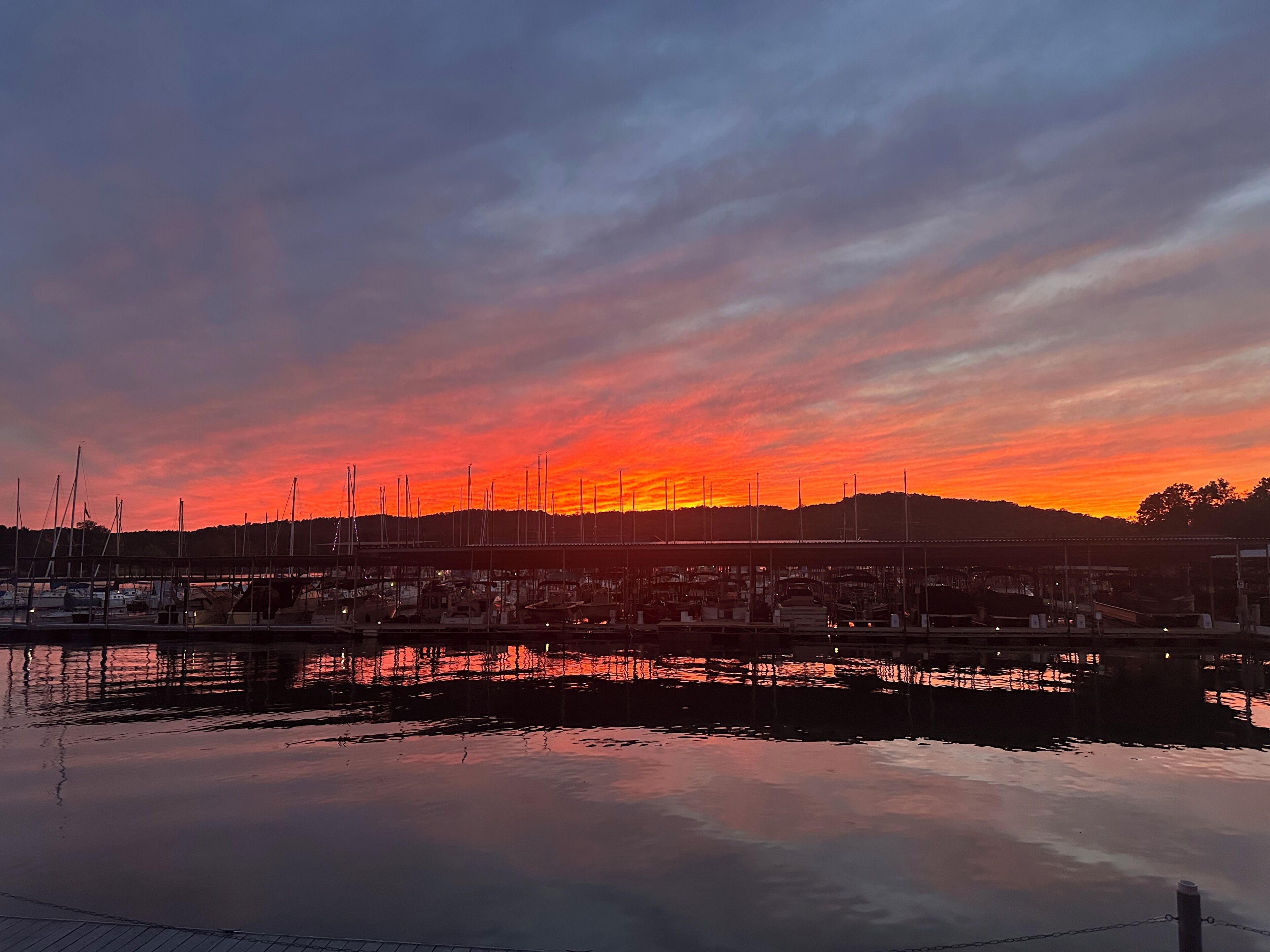 Eine Marina mit vertäuten Booten, eine ruhige Wasseroberfläche, die den Himmel spiegelt, und eine hügelige Landschaft im Hintergrund.