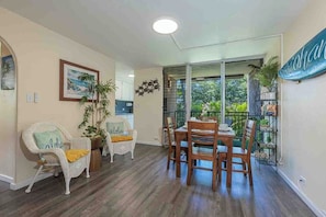 A dining area with a wooden table and chairs, a wicker chair with a cushion, and a potted plant. - Kalama Terrace L203 South Kihei Retreat with Pool, Lanai, Ocean Nearby (Kihei)