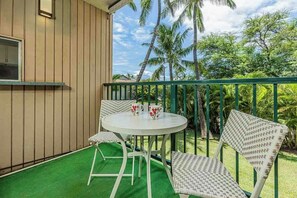 A balcony with a white table and wicker chairs, overlooking a tropical garden with palm trees. - Kalama Terrace L203 South Kihei Retreat with Pool, Lanai, Ocean Nearby (Kihei)