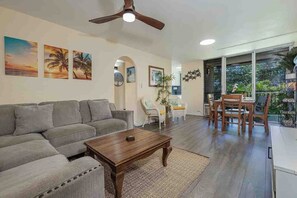 A living room with a sofa, coffee table, and a dining area with chairs. There are ceiling fans, a large window, and artwork on the walls. - Kalama Terrace L203 South Kihei Retreat with Pool, Lanai, Ocean Nearby (Kihei)