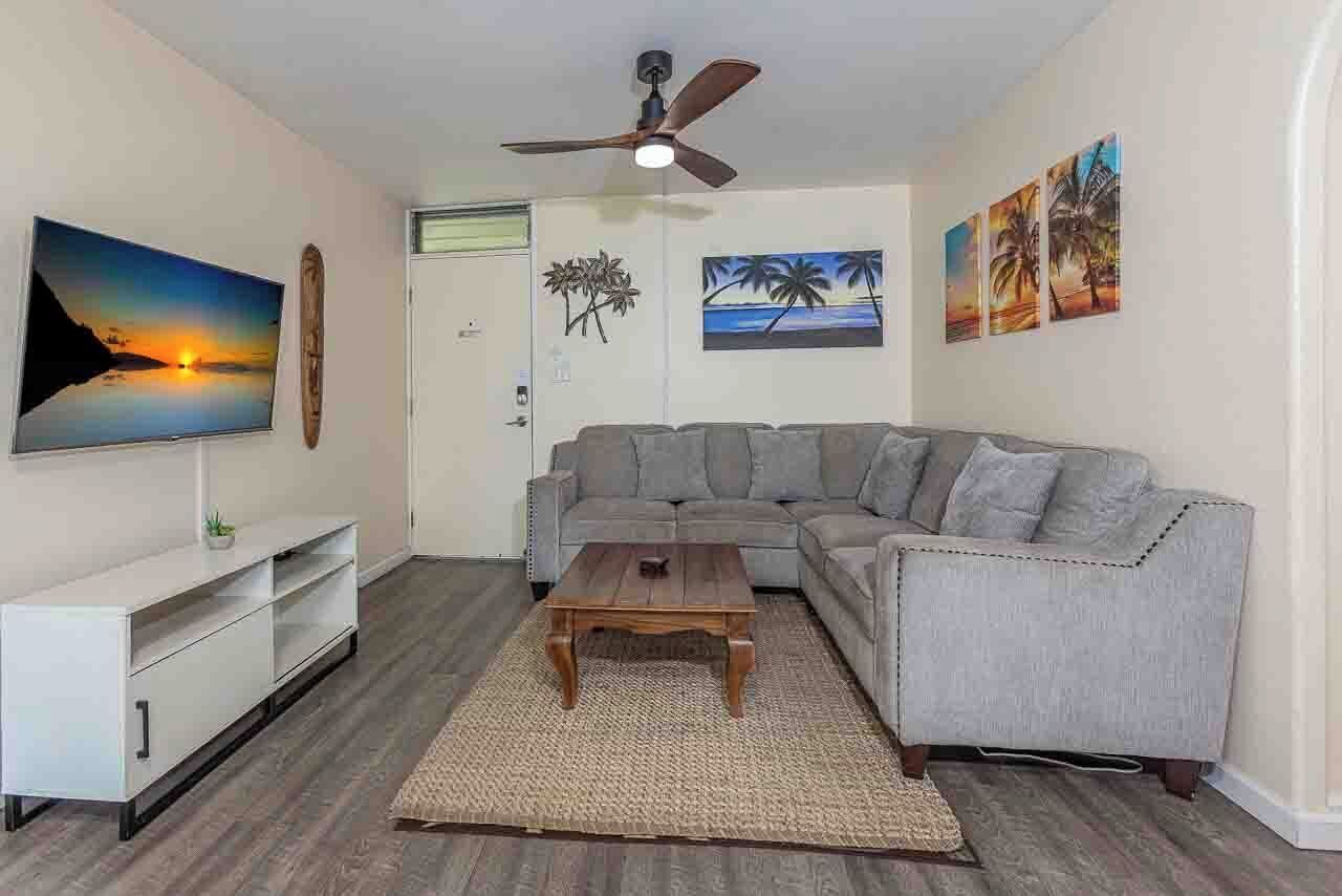 A living room with a grey sofa, a wooden coffee table, a flat-screen TV, and wall art depicting tropical scenes.