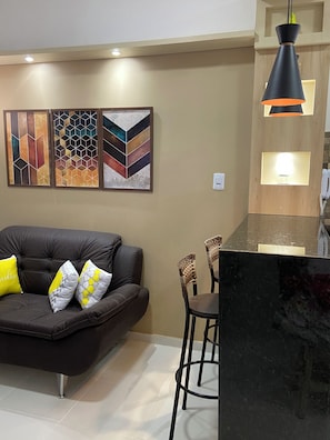 A modern living room with a dark leather sofa, two chairs, and a coffee table. There are three abstract paintings on the wall and a pendant light above the table. - 207 Apto 2 Qtos Lindo (Belém)