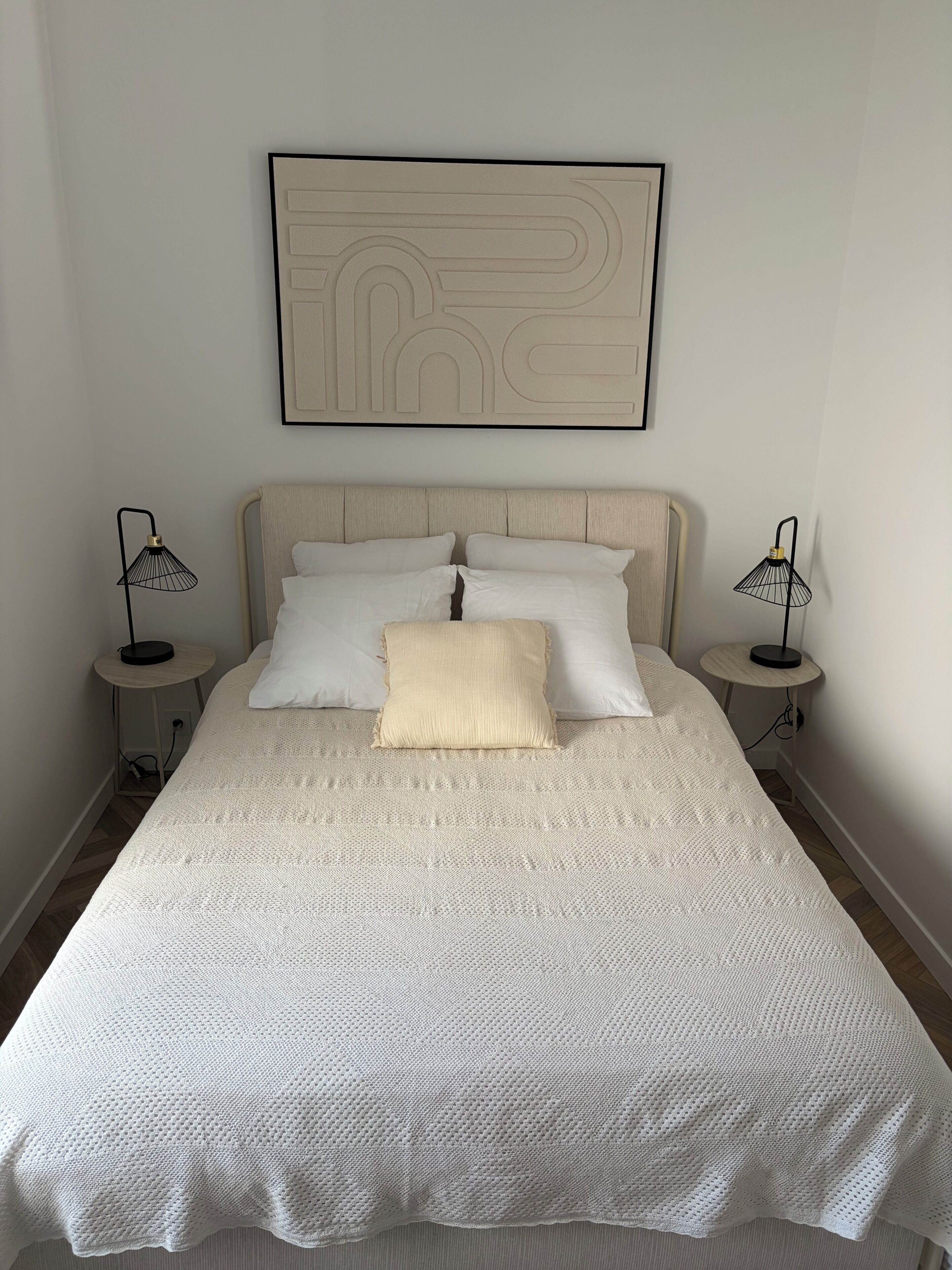 A neatly made bed with a textured white duvet, two bedside lamps, and a wall-mounted abstract art piece.