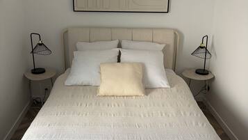 A neatly made bed with a textured white duvet, two bedside lamps, and a wall-mounted abstract art piece.