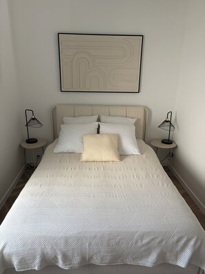 Uma cama bem arrumada com um edredom branco texturizado, duas luminárias de mesa e uma obra de arte abstrata na parede.