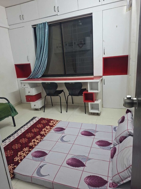 A bedroom with a bed, a desk with two chairs, a window with a curtain, and a wardrobe.