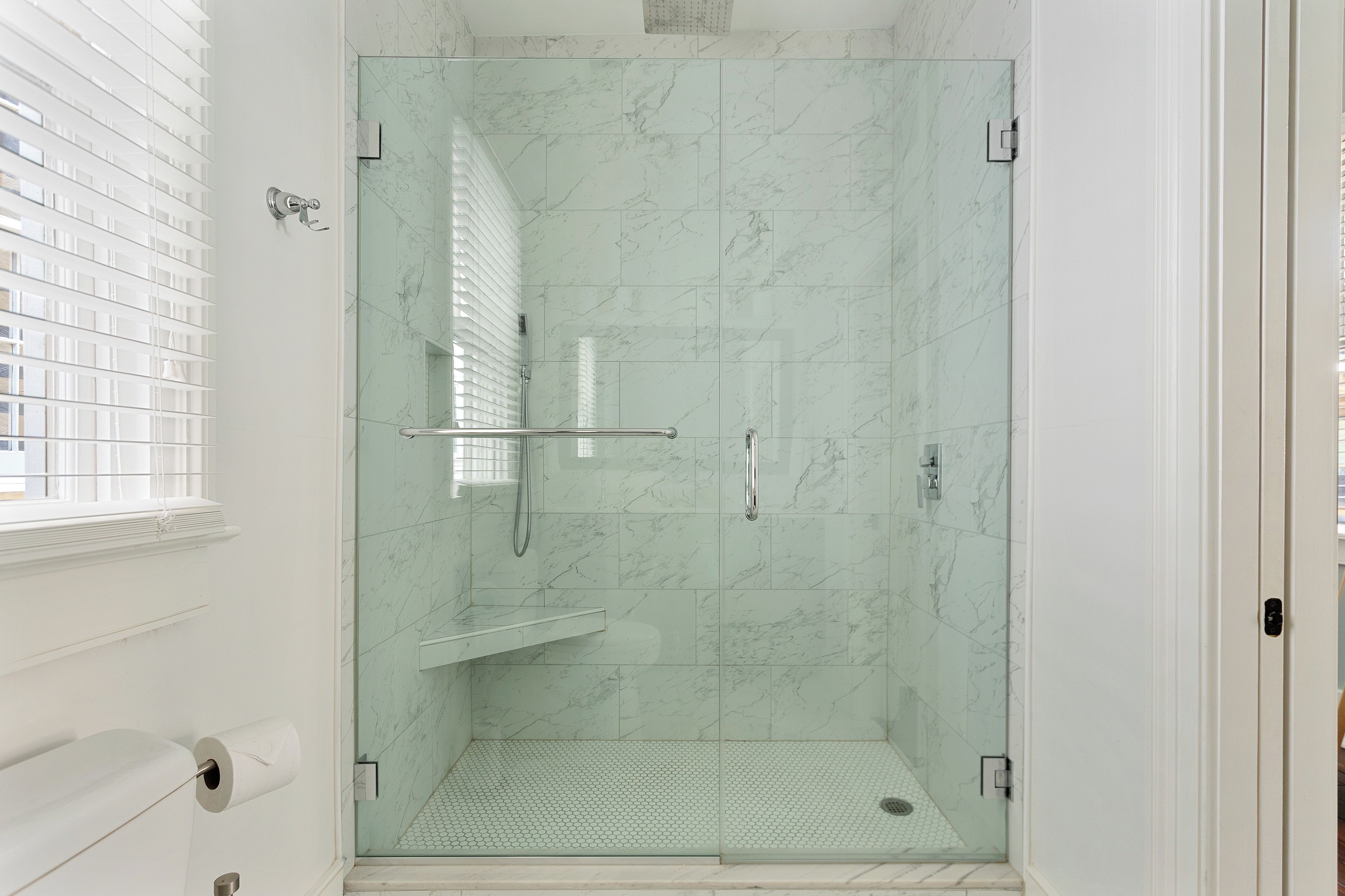 A glass-enclosed shower with a handrail, a tiled floor, and a view of a building outside.