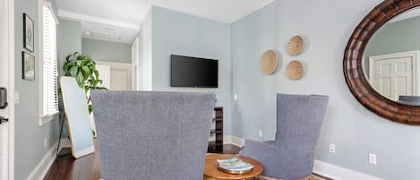 A living room with two grey armchairs, a round mirror, a flat-screen TV, and a small wooden table with a magazine.