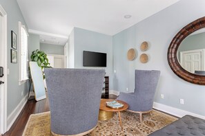 A living room with two grey armchairs, a round mirror, a flat-screen TV, and a small wooden table with a magazine. - Smalls Alley (Charleston)