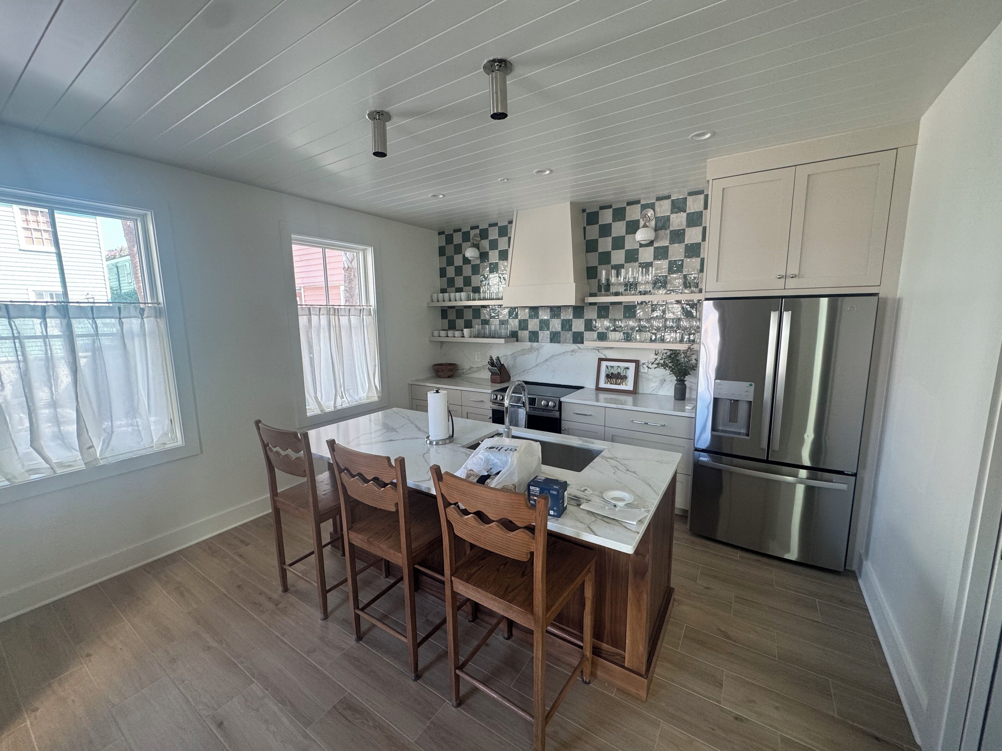 A kitchen with a marble countertop, wooden chairs, a stainless steel refrigerator, and a window with curtains.
