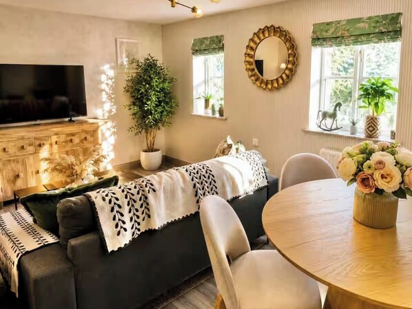 Ein Wohnzimmer mit einer Couch, einem Fernseher, einem Esstisch mit Stühlen, einer Topfpflanze und einem Spiegel an der Wand.