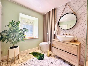 A bathroom with a large potted plant, a toilet, a sink with a mirror, and a window. - Muddy Boots Arcade Royale (Kirkby Stephen)