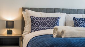 Un letto rifatto con cura, con una testiera grigia, cuscini a motivi bianchi e blu e una lampada da comodino su un tavolino.