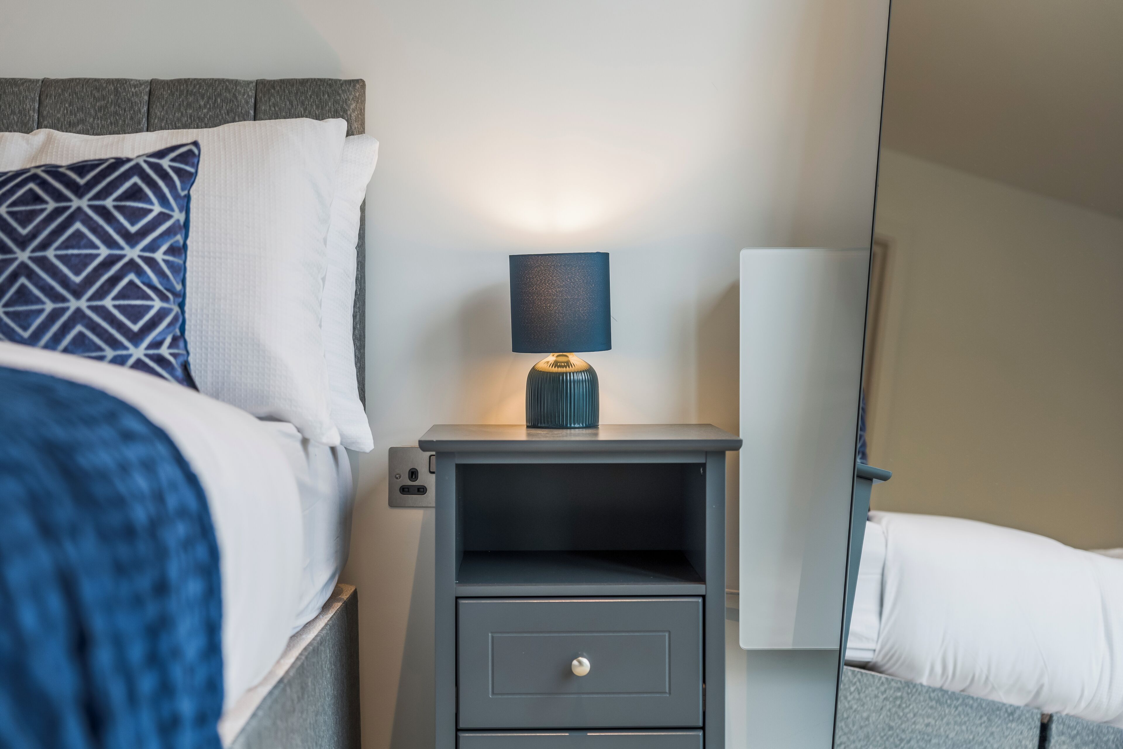 A neatly made bed with a patterned pillow, a bedside table with a lamp, and a drawer.