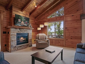 A cozy cabin interior with a stone fireplace, a comfortable armchair, a wooden coffee table, and a large window offering a view of the outdoors.