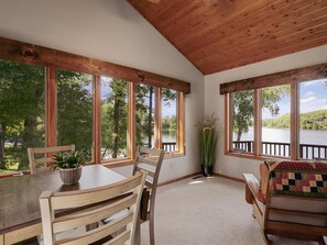 A room with a wooden ceiling, large windows overlooking a lake, a dining table with chairs, and a cozy seating area with a quilted cushion.