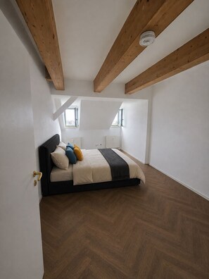 Quarto com cama, vigas de madeira, paredes brancas e padrão de piso em espinha de peixe.