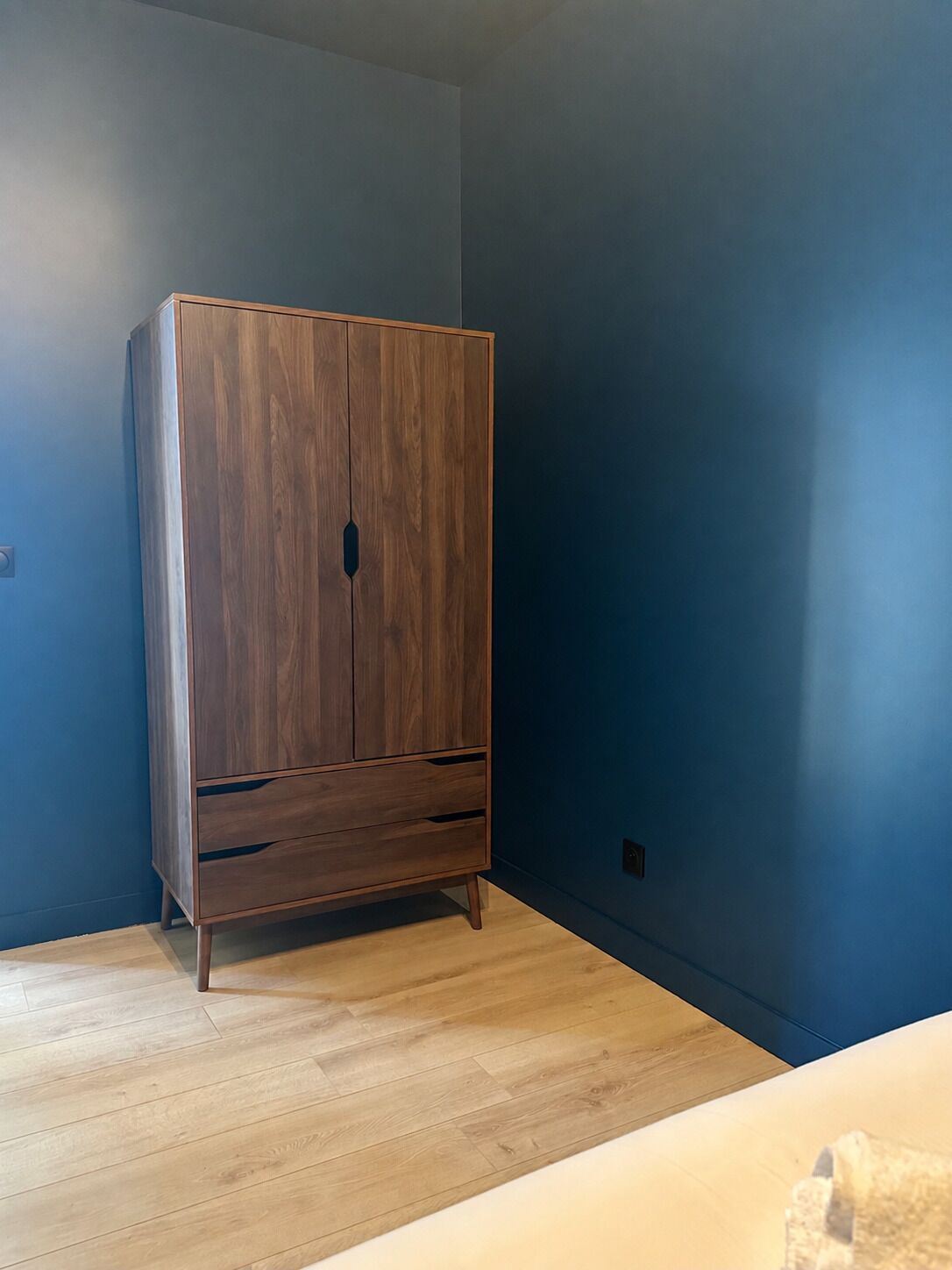 A room with a wooden wardrobe against a blue wall, wooden flooring, and a visible electrical outlet.