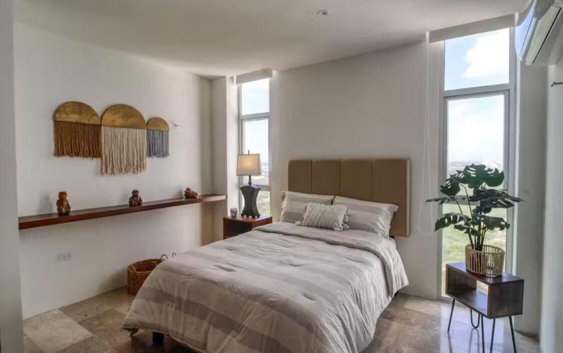A bedroom with a large bed, a nightstand, a lamp, a wall-mounted shelf with decorative items, and a window with a view.