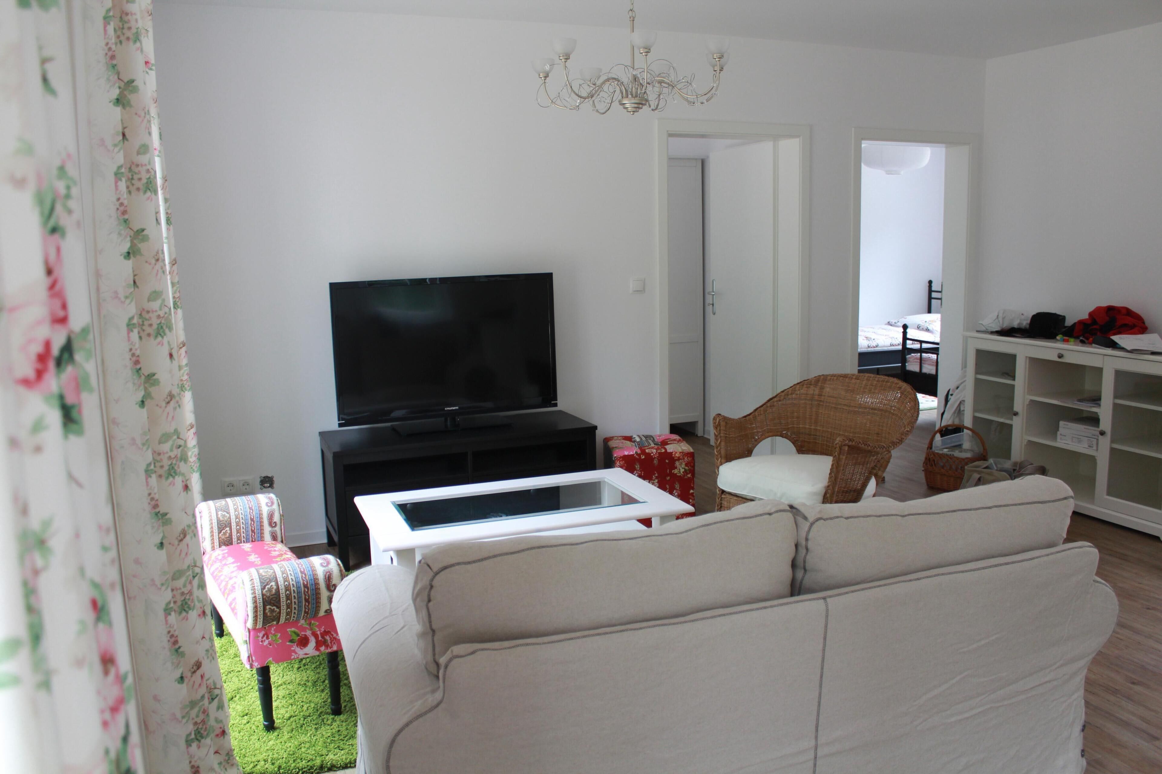 Ein Wohnzimmer mit Flachbildfernseher, einer Couch, einem Korbsessel und einer weißen Kommode.