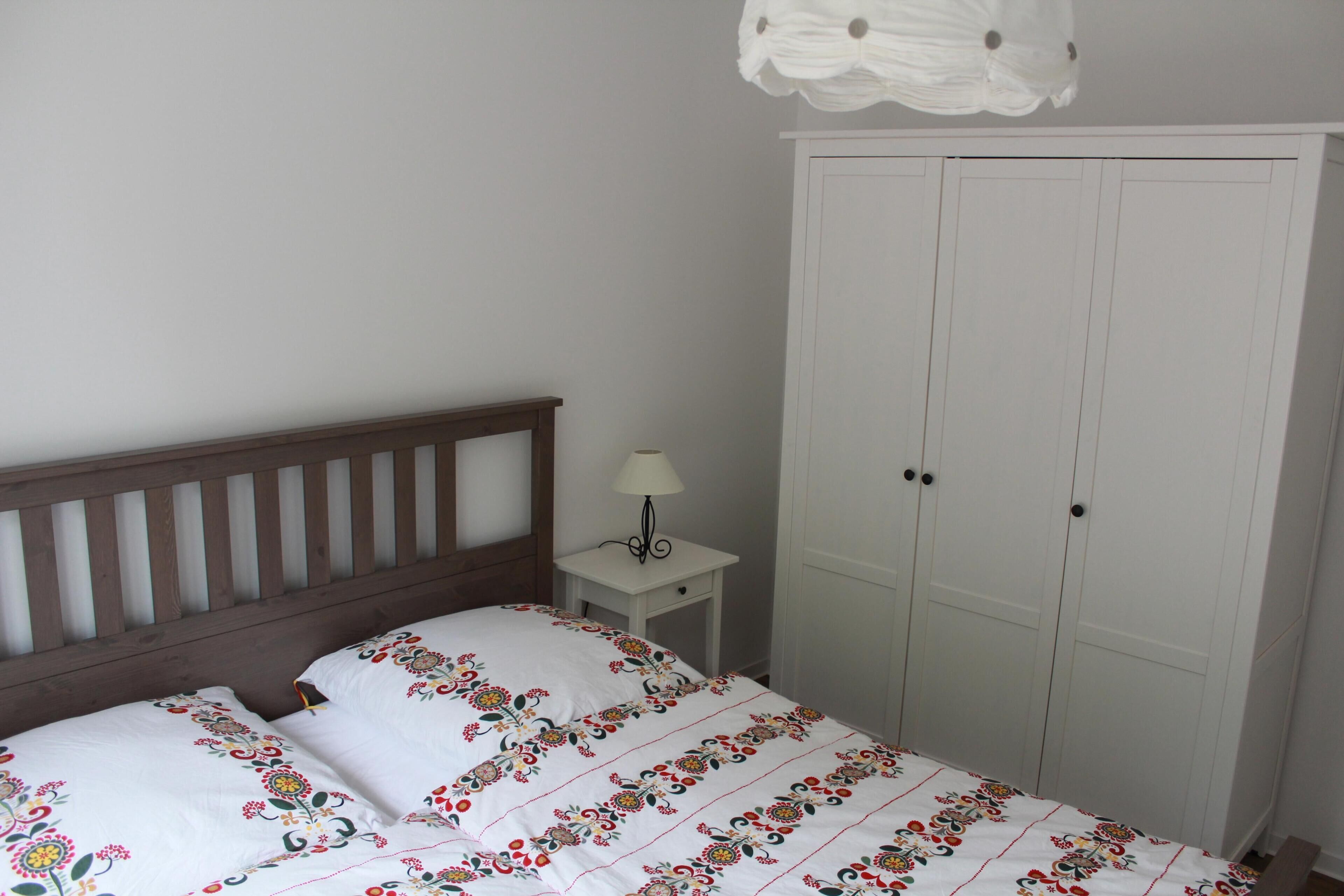 Ein Schlafzimmer mit einem Holz-Kopfteil, einem weißen Bett mit floralem Bettzeug, einem Nachttisch mit Lampe und einem weißen Kleiderschrank.